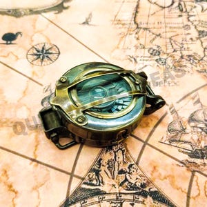 以下が含まれることがあります： ガラスレンズを備えたヴィンテージ真鍮コンパス。古い地図を背景に配置。コンパスは、ヒンジ付きの蓋と方向ダイヤルを備えた詳細なデザイン。地図は色あせた色と航海のイラストが特徴です。