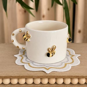 May include: A white ceramic mug featuring three-dimensional gold and black bee accents on the exterior and handle. The handle is decorated with gold polka dots. The mug sits on a white scalloped fabric coaster with gold and silver embroidered trim.