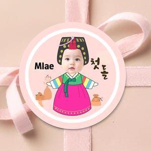 May include: A round sticker with a baby wearing a traditional Korean hanbok. The sticker has the word "Mlae" and Korean text. The baby is wearing a pink skirt, a green top, and a black hat with gold accents. The sticker is on a pink background.