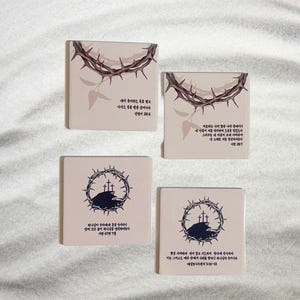 May include: Four beige square coasters with black and white illustrations of thorns, crosses, and a hill. Each coaster has a different bible verse printed in black text. The verses are: "I thirst" John 19:28, "He was pierced for our transgressions, he was crushed for our iniquities; the punishment that brought us peace was upon him, and by his wounds we are healed." Isaiah 53:5, "And he said to them, "I tell you the truth, some who are standing here will not taste death before they see the Son of Man coming in his kingdom." Matthew 16:28, "And he said to them, "I tell you the truth, some who are standing here will not taste death before they see the Son of Man coming in his kingdom." Matthew 16:28