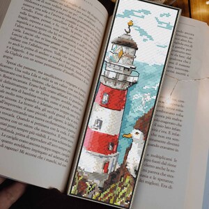 Könnte beinhalten: Ein rot-weißer Leuchtturm mit einer Möwe, die auf einem Felsen sitzt. Der Leuchtturm ist von blauem Wasser und einem bewölkten Himmel umgeben. Das Bild ist ein Kreuzstichmuster.