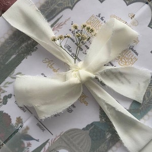 May include: A white ribbon tied in a bow with a small bouquet of white flowers on top. The ribbon is tied around a folded card with a floral design and the text "This occasion marks..." and "Abraham Khan".