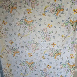 Peut inclure: Un tissu blanc avec un motif répété de lapins, de fleurs et de papillons. Les lapins tiennent un arc-en-ciel et sont entourés de fleurs roses, violettes et jaunes. Les papillons sont jaunes avec des accents orange.