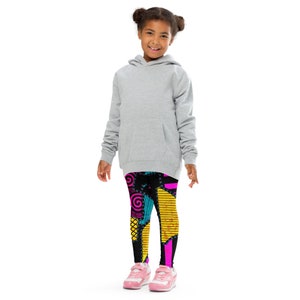May include: A young person wearing a gray hoodie and black leggings with a colorful patchwork design. The leggings feature a variety of shapes, including circles, stars, and stripes, in shades of pink, yellow, blue, and black.