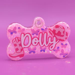 Könnte beinhalten: Rosaer Knochen-Hundeanhänger mit Glitzer, mit einem rosa Cowboyhut, einem rosa Cowboystiefel und rosa Schmetterlingen. Der Anhänger ist mit dem Namen "Dolly" personalisiert.