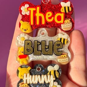 May include: Three bone-shaped pet tags with names and cartoon character designs. The top tag is red glitter with the name "Thea" in yellow. The middle tag is clear with "Blue" in gold. The bottom tag is black glitter with "Hunny".