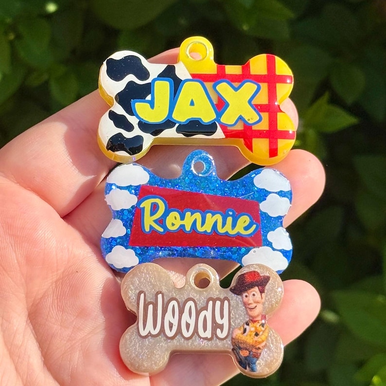Puede incluir: Tres etiquetas para mascotas con forma de hueso. La etiqueta superior tiene un estampado de vaca en blanco y negro y el nombre "JAX". La etiqueta del medio es azul con el nombre "Ronnie". La etiqueta inferior es de color beige con el nombre "Woody" y una imagen de dibujos animados.