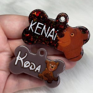 Pode incluir: Duas etiquetas para cães em forma de osso com glitter e imagens de ursos de desenho animado. A etiqueta superior é vermelha e preta com glitter e diz "KENAI" com uma imagem de urso de desenho animado. A etiqueta inferior é marrom com uma imagem de urso de desenho animado e diz "KODA".