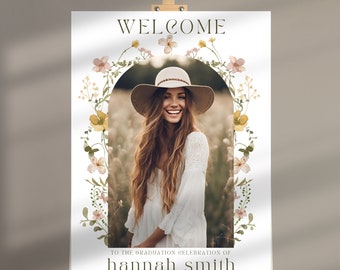 Può includere: Benvenuti alla celebrazione di laurea di hannah smith. Un cartello bianco con un bordo a forma di corona di fiori e una foto di una donna con un vestito e un cappello bianchi. Il cartello è su un cavalletto.