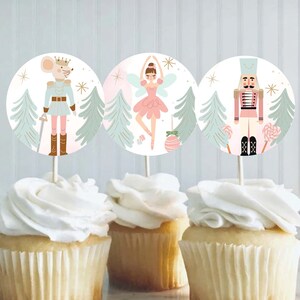 May include: Three cupcakes topped with white frosting and decorated with round, colorful paper toppers featuring a nutcracker, a ballerina, and a mouse in a soldier's uniform. The toppers have a light blue background with green trees and gold stars.