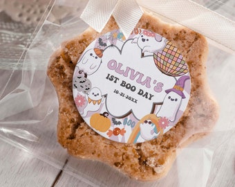 May include: A star-shaped sugar cookie decorated with a round sticker featuring a ghost theme. The sticker says "Olivia's 1st Boo Day 10-31-20XX".