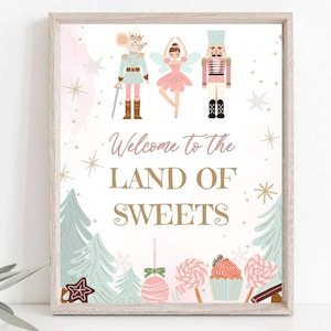 Puede incluir: Un cartel blanco con un fondo de acuarela rosa y azul. El cartel dice "Bienvenido a la tierra de los dulces" en letras doradas. Hay ilustraciones de un ratón con un disfraz de cascanueces, una bailarina y un soldado cascanueces.