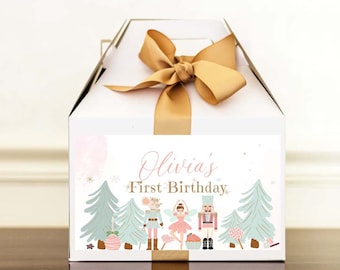 May include: A white gift box with a gold ribbon and a label that says "Olivia's First Birthday". The label features a pink and green illustration of a ballerina, a nutcracker, and a mouse in a forest setting.