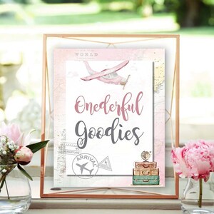 May include: A pink and gray sign with the text "Onederful Goodies" on a white background. The sign is decorated with a pink airplane, a globe, and vintage suitcases. The background is a pink and white world map.