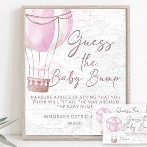 May include: Pink and white hot air balloon themed baby shower game. Guests measure a piece of string to guess the size of the mom-to-be's baby bump. The game card says "Guess the Baby Bump" and includes instructions to wrap the string around the card and sign your name.