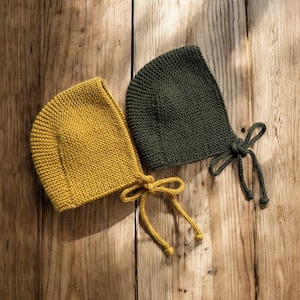 May include: Two knitted baby bonnets. One is mustard yellow, the other is dark green. Both have a rounded shape and tie closures. The bonnets are displayed on a rustic wooden surface.