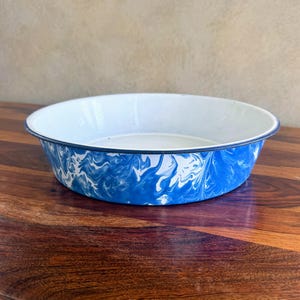 May include: A round, enamelware bowl with a white interior and a blue and white swirled exterior pattern. The rim is dark blue. The bowl sits on a wooden surface.