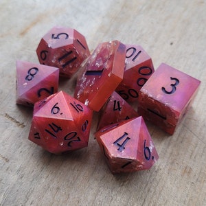 May include: A collection of seven multi-sided dice in red and pink hues, with silver inclusions and black numerals. The dice are crafted from a translucent material, featuring various shapes, such as a d20, d12, d10, d8, d6, and d4.