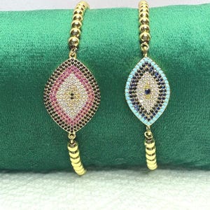 May include: Two gold-colored beaded bracelets. Each bracelet features a dangling, eye-shaped charm. One charm has pink, white, and black stones. The other charm has blue, white, and black stones. Both charms have a central gold-colored diamond shape.