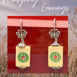 May include: Mahjong Earrings. Silver-toned earrings with a butterfly accent and a rectangular, cream-colored tile featuring a red and green circular design. The text "Mahjong Earrings" is written in cursive.