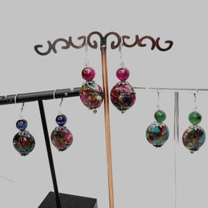 May include: Earrings displayed on a decorative stand. The earrings feature a variety of colorful beads, including pink, blue, green, and multi-colored spherical and round beads. The earrings are suspended from silver-colored hooks.