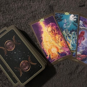 May include: A set of tarot cards with a dark green border and a black background with gold stars. The cards feature a triple moon symbol with a pentagram inside. The cards are arranged in a fan shape with the top card showing a woman holding a glowing orb.