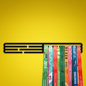 May include: Black metal medal display rack with multiple colorful race ribbons hanging on it. The ribbons have text on them, including "21,0975 km", "SIGULDAS MARATONS", "RĒZEKNES PUSMARATONS", "WERUN RIGA", "JA POOLMARATON", "17.05.2015 LATTELE", "K", "PUSMARATONS 21,0975 km", "ARKA NS", "2015", "PUSM", "PIEDZĪVOJUMS".