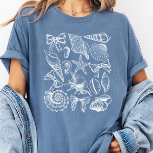 May include: A blue t-shirt featuring a white illustration of various seashells, starfish, and bows. The shirt has rolled-up sleeves and is paired with a denim jacket and shorts. The overall theme is coastal and beach-inspired.