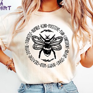 May include: Cream-colored t-shirt with a black bee graphic inside a circular design. The design includes words like "Kind," "Positive," and "Creative." The text "NEW ERA TEESHIRT" is printed at the top left.