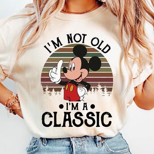 Puede incluir: Camiseta color crema con Mickey Mouse y el texto "I'M NOT OLD I'M A CLASSIC". Mickey Mouse levanta el dedo, sobre un dise&ntilde;o de puesta de sol. La camiseta se combina con vaqueros azul claro.