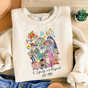 May include: Cream-colored sweatshirt featuring a colorful graphic of Toy Story characters. The design includes Woody, Buzz Lightyear, and others, with the text "Toy Story Land" and "To Infinity and Beyond! Est. 1995". The sweatshirt is paired with blue denim shorts.