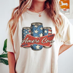 May include: A white t-shirt with a vintage-style graphic of three red, white, and blue American flag-themed cans with the text "Ameri Can" in red.