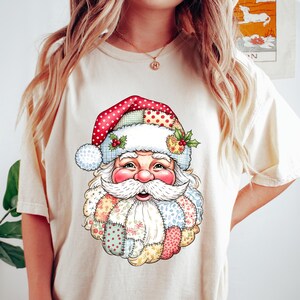 May include: Cream-colored t-shirt featuring a colorful Santa Claus graphic. The Santa design includes a red and white polka-dot hat, a white beard, and a patchwork-style coat. The shirt is a casual, comfortable style.