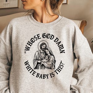 May include: A light gray sweatshirt with a black graphic of a religious family and the text "WHOSE GOD DAMN WHITE BABY IS THIS?" in a circular design. The sweatshirt is a casual style.