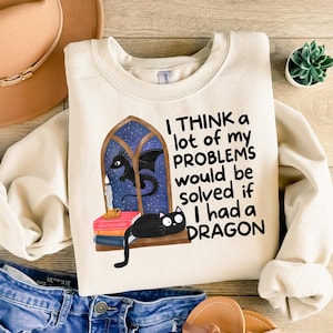 May include: Cream-colored sweatshirt with a whimsical design. It features a black cat, a dragon, and the text "I THINK a lot of my PROBLEMS would be solved if I had a DRAGON." The design includes a window, books, and a candle.