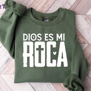 May include: A dark green sweatshirt with the text "DIOS ES MI ROCA" in white block letters. The word "ROCA" features a cross in place of the letter "O" and a heart symbol. The words "NEW ERA TEESHIRT" are in the upper left corner.