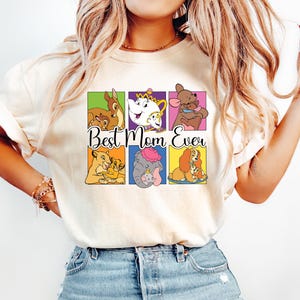 May include: Cream-colored t-shirt featuring a colorful graphic of Disney characters in square frames. The phrase "Best Mom Ever" is written in a script font below the images. The shirt is paired with light blue jeans.