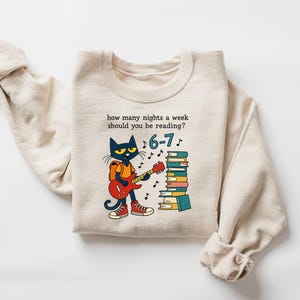 May include: Cream-colored sweatshirt featuring a cartoon cat playing a red guitar next to a stack of books. The text reads, "how many nights a week should you be reading?" with the answer "6-7".