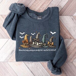 May include: A dark gray sweatshirt featuring a graphic of several witch hats in various colors, including orange, brown, and green, with white bats. The text "Wicked but pretty, spooky got sparkly, that's my kind of witchcraft" is printed below the hats.