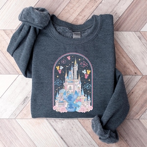 K&ouml;nnte beinhalten: Dunkelgraues Sweatshirt mit einem farbenfrohen Aufdruck eines Schlosses, Feuerwerk, Ballons und einer Zeichentrickfigur. Die Grafik ist von einem rosa Bogen umrahmt und von rosa Blumen umgeben. Das Sweatshirt ist aus weichem Material.