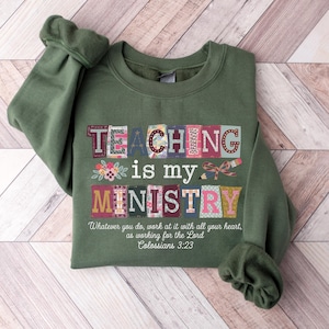 May include: An olive green sweatshirt with the words "TEACHING is my MINISTRY" in a colorful, patchwork-style design. The text "Whatever you do, work at it with all your heart, as working for the Lord Colossians 3:23" is printed below.