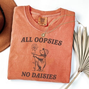 May include: A burnt orange t-shirt with the text "ALL OOPSIES NO DAISIES" printed on it. A cartoon bear holding a flower is printed above the text.