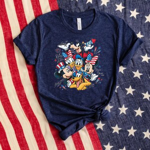 Może przedstawiać: Granatowa koszulka z kr&oacute;tkim rękawem z nadrukiem graficznym przedstawiającym Myszkę Miki, Myszkę Minnie, Kaczora Donalda, Daisy, Goofy'ego i Pluto w patriotycznych czerwono-biało-niebieskich akcesoriach, w otoczeniu ilustracji fajerwerk&oacute;w i gwiazd.