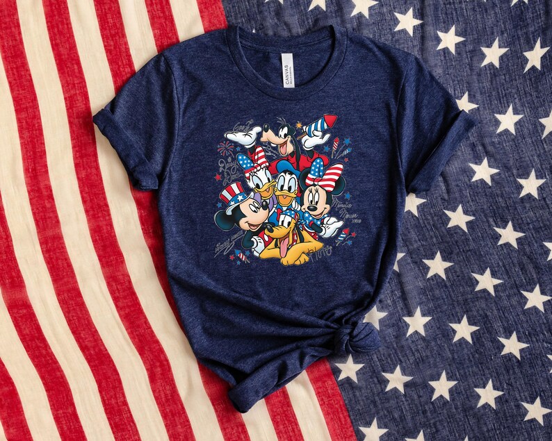 Może przedstawiać: Granatowa koszulka z kr&oacute;tkim rękawem z nadrukiem graficznym przedstawiającym Myszkę Miki, Myszkę Minnie, Kaczora Donalda, Daisy, Goofy'ego i Pluto w patriotycznych czerwono-biało-niebieskich akcesoriach, w otoczeniu ilustracji fajerwerk&oacute;w i gwiazd.