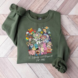 May include: An olive green sweatshirt featuring a colorful graphic of characters from the animated film Toy Story. The design includes Woody, Buzz Lightyear, and other characters, with the text "Toy Story Land" and "To Infinity and Beyond! Est 1995".