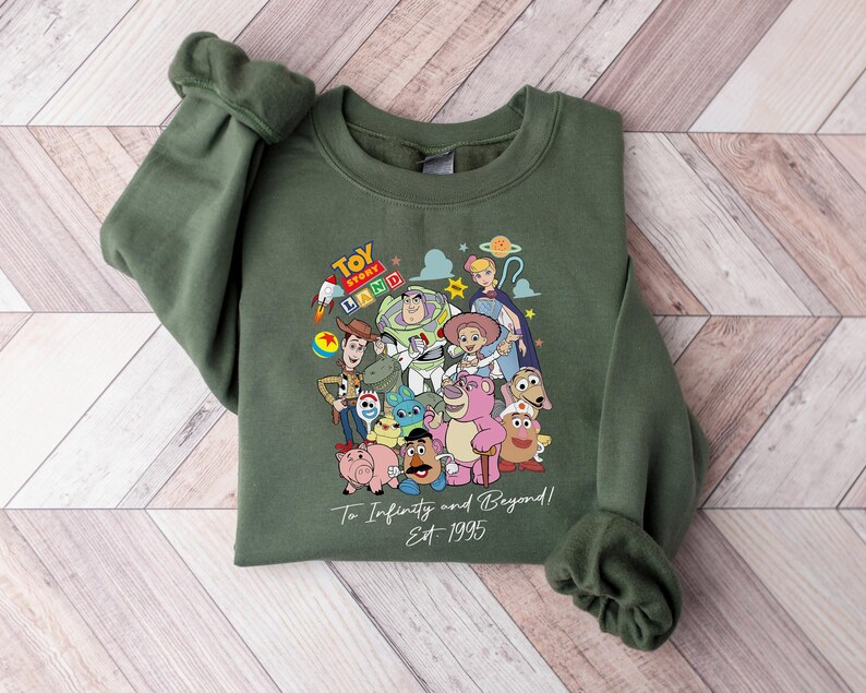 May include: An olive green sweatshirt featuring a colorful graphic of characters from the animated film Toy Story. The design includes Woody, Buzz Lightyear, and other characters, with the text "Toy Story Land" and "To Infinity and Beyond! Est 1995".
