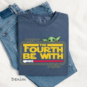 May include: A navy blue t-shirt featuring the text "MAY THE FOURTH BE WITH YOU" in yellow, with a green character peeking over the text. A red lightsaber graphic is below the text. The shirt is folded on top of a pair of blue jeans.