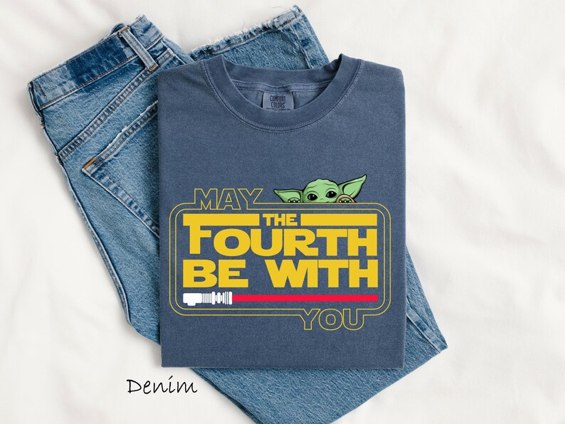 May include: A navy blue t-shirt featuring the text "MAY THE FOURTH BE WITH YOU" in yellow, with a green character peeking over the text. A red lightsaber graphic is below the text. The shirt is folded on top of a pair of blue jeans.