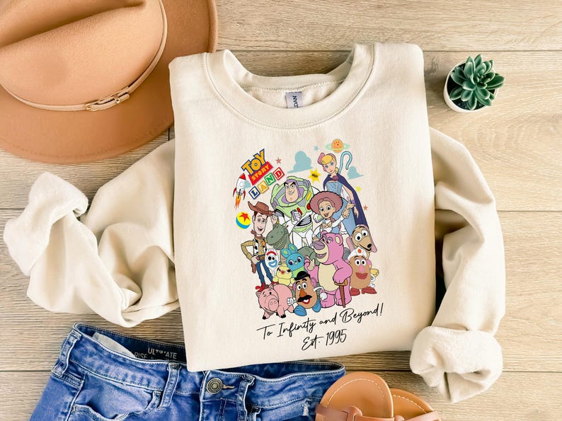 May include: Cream-colored sweatshirt featuring a colorful graphic of Toy Story characters. The design includes Woody, Buzz Lightyear, and others, with the text "Toy Story Land" and "To Infinity and Beyond! Est. 1995". The sweatshirt is paired with blue denim shorts.