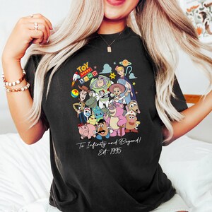 May include: Black t-shirt featuring a colorful graphic of Toy Story characters, including Woody, Buzz Lightyear, and others. The shirt has the text "Toy Story Land" at the top and "To Infinity and Beyond! Est 1995" below the characters.